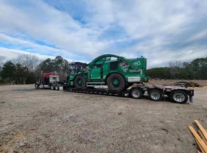 JW Hart Transport semi hauling a large green forestry chipper on a lowboy trailer