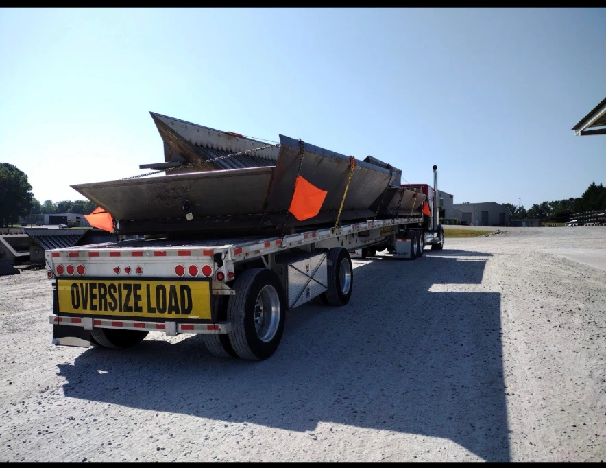 Oversize load flatbed trailer hauling chained steel equipment on a sunny gravel lot