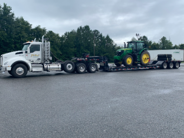 White JW Hart Transport semi hauling a green John Deere tractor on a lowboy trailer