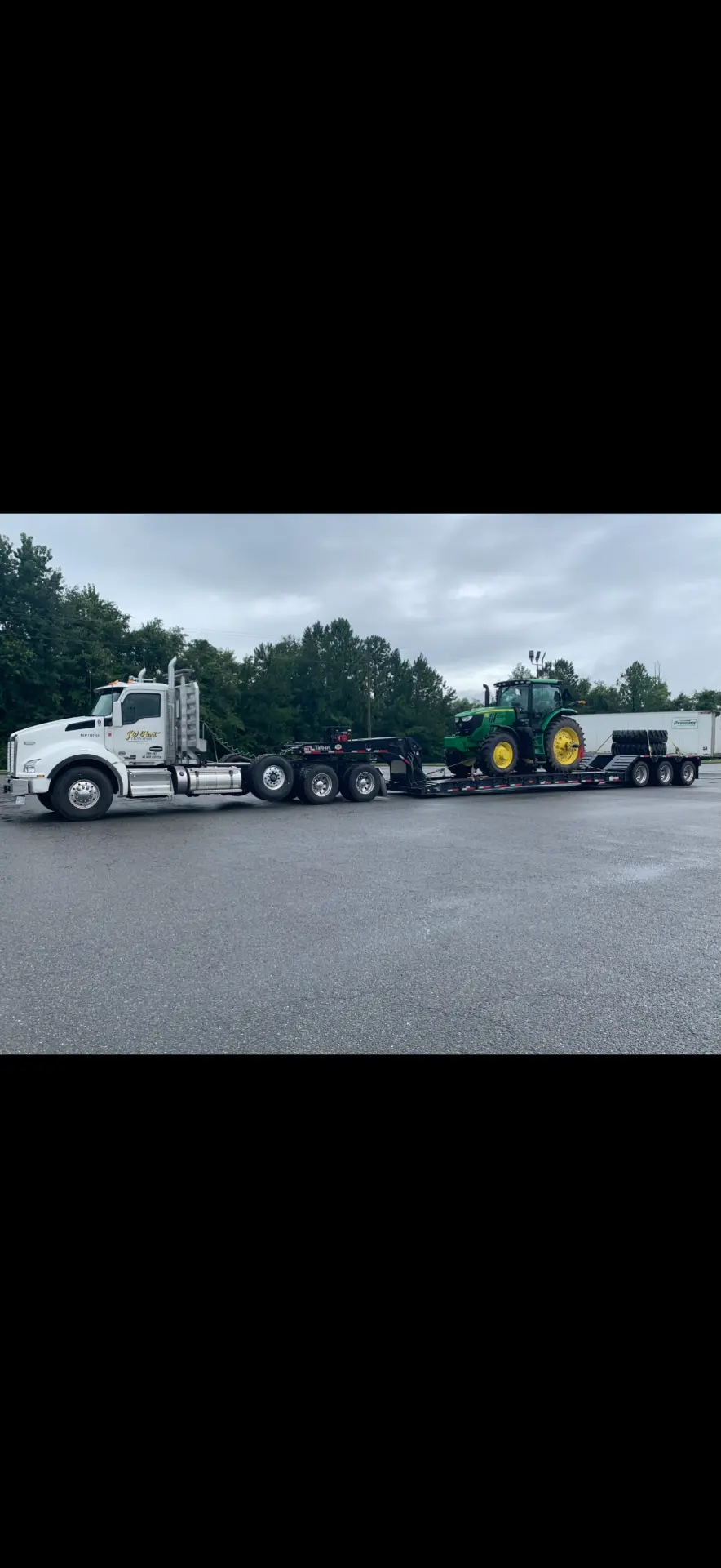White JW Hart Transport semi hauling a green John Deere tractor on a lowboy trailer