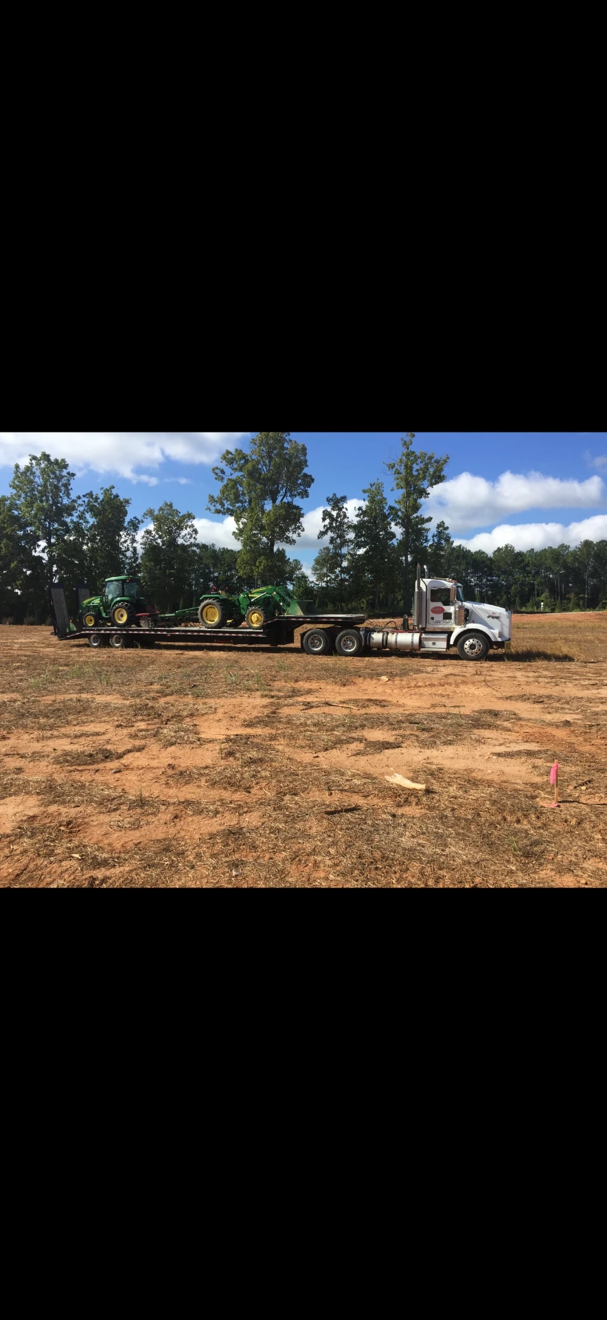 White JW Hart Transport semi hauling two green John Deere tractors at a rural jobsite