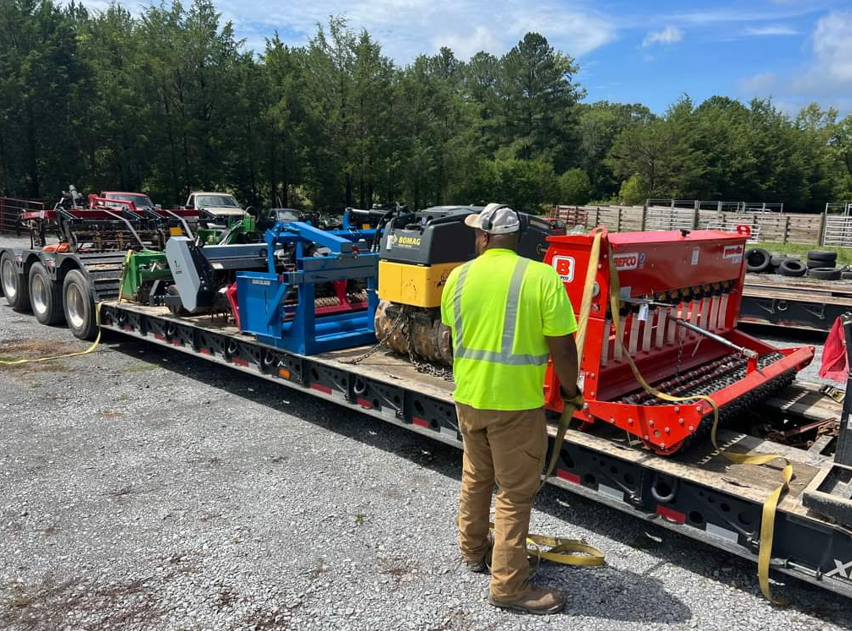 Worker securing a Bobcat, blue equipment, and red seeder on a JW Hart Transport lowboy trailer